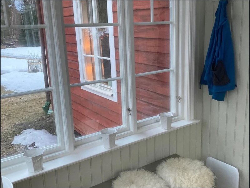 Fönsterraden inifrån glasverandan, med vita spöjsade bågar, blomkrukor i fönsterkarmen och sittbänk under
