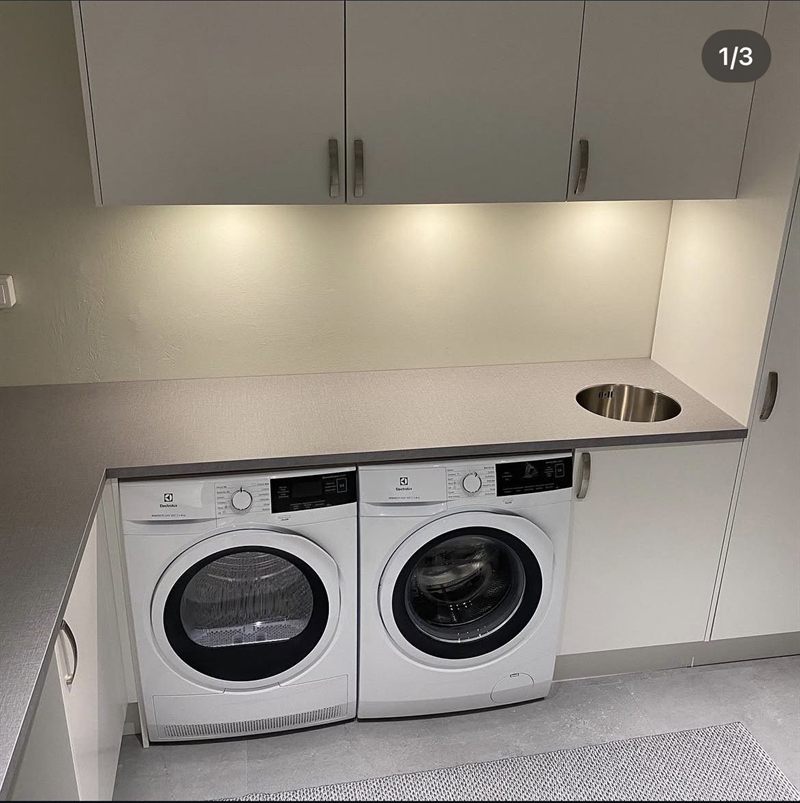 Tvättstuga med tvättmaskin och torktumlare inbyggda under bänkskiva, överskåp och rund tvättho